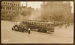 La foule a disparu, laissant place à des gens à l'arrière-plan. Le tramway, qui avait été renversé, est maintenant debout. De la fumée s'échappe du tramway. Une automobile et trois hommes se tiennent à côté.