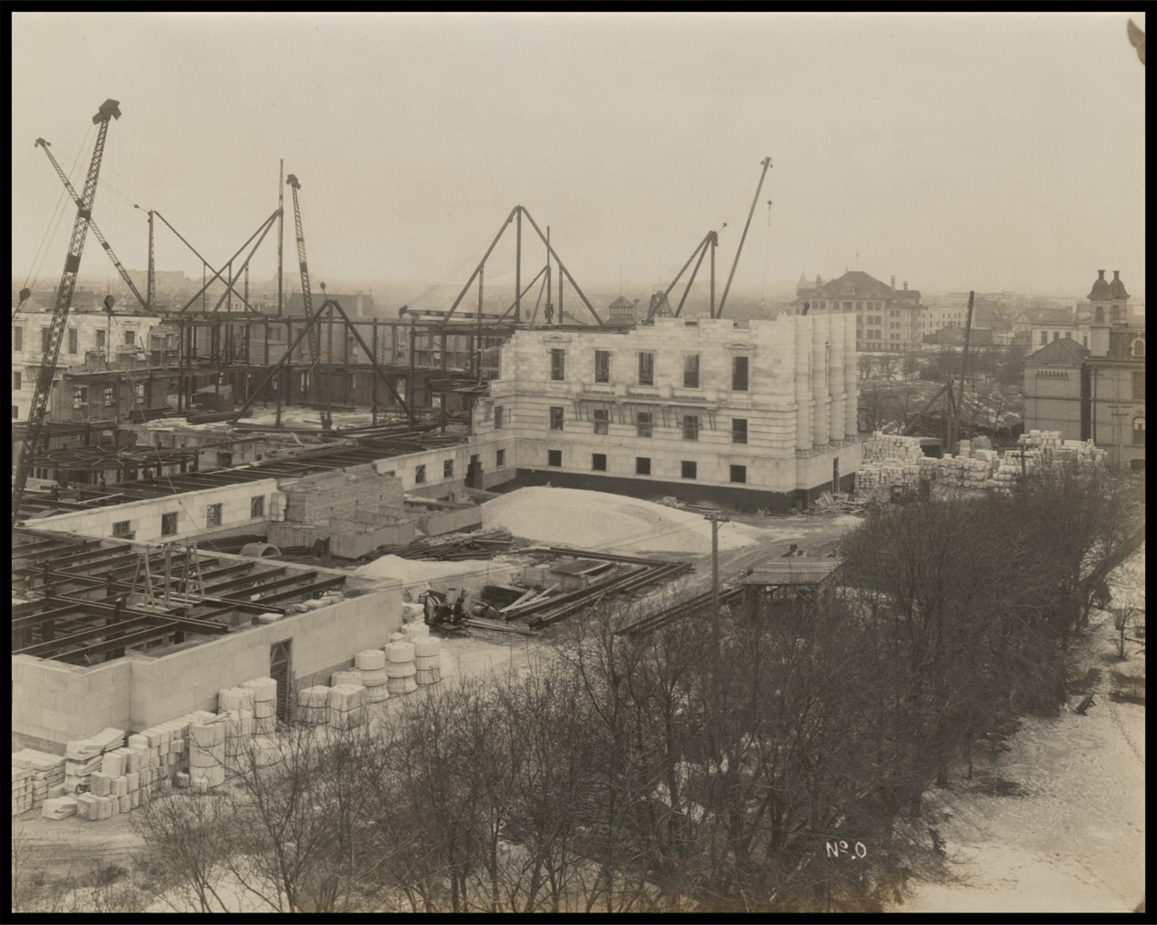 Photo with aerial view of the east side of the Legislative Building under construction. Some walls are built, while others are just steel frames. There is no roof. Large cranes and construction materials surround the building.