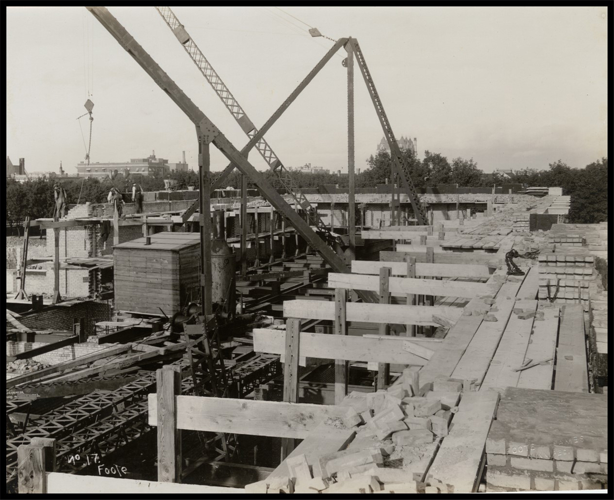 Photo of the construction from the third floor, which is comprised of incomplete wood planks. Large cranes and steel frames are in the middle of the building. There are stacks of bricks in the forground, while masons lay bricks along the back wall.