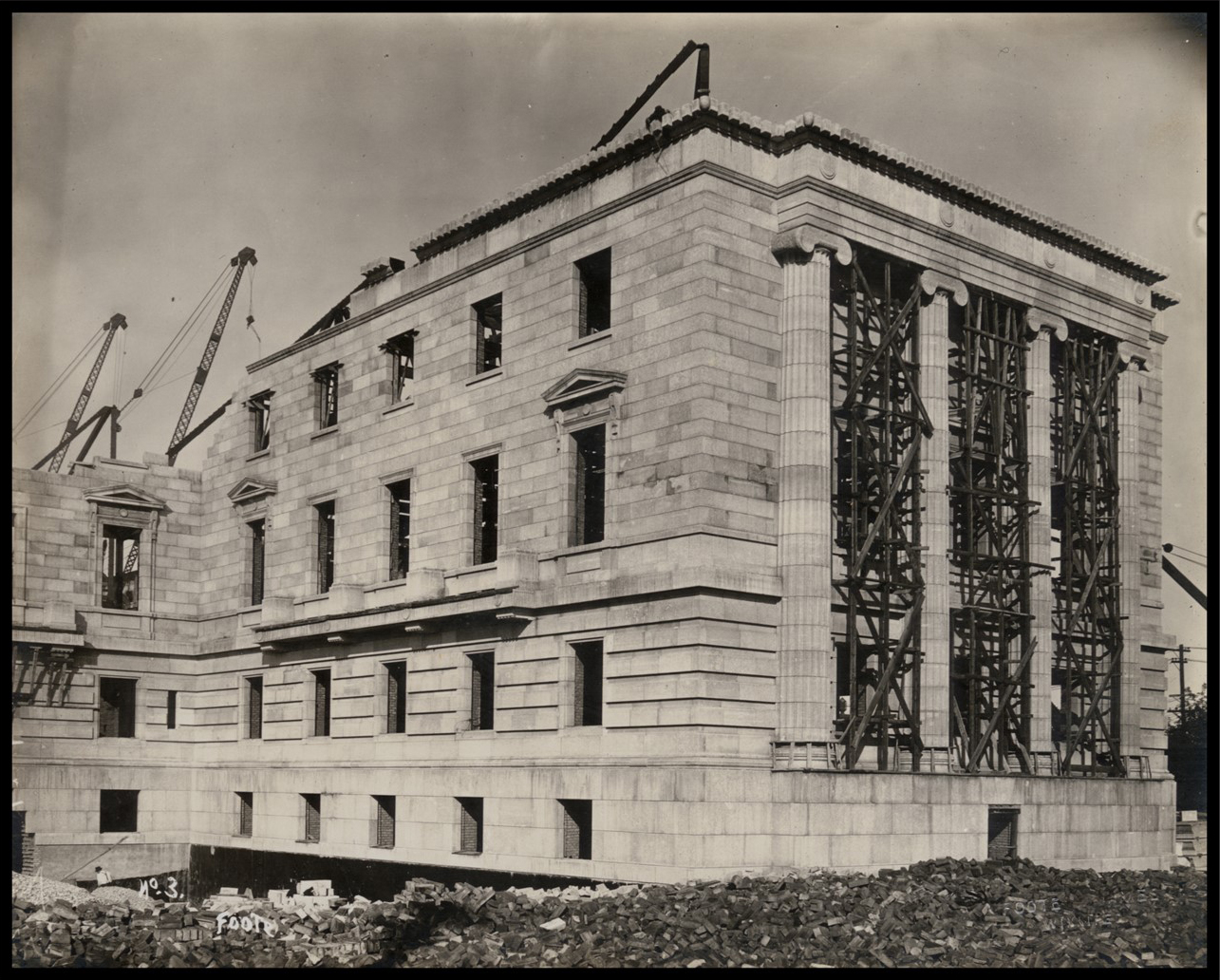 Photo of the exterior of an almost complete wall. There are no windows. There is scaffolding between the stone columns. In the background, giant cranes are visible through incomplete portions of the wall.
