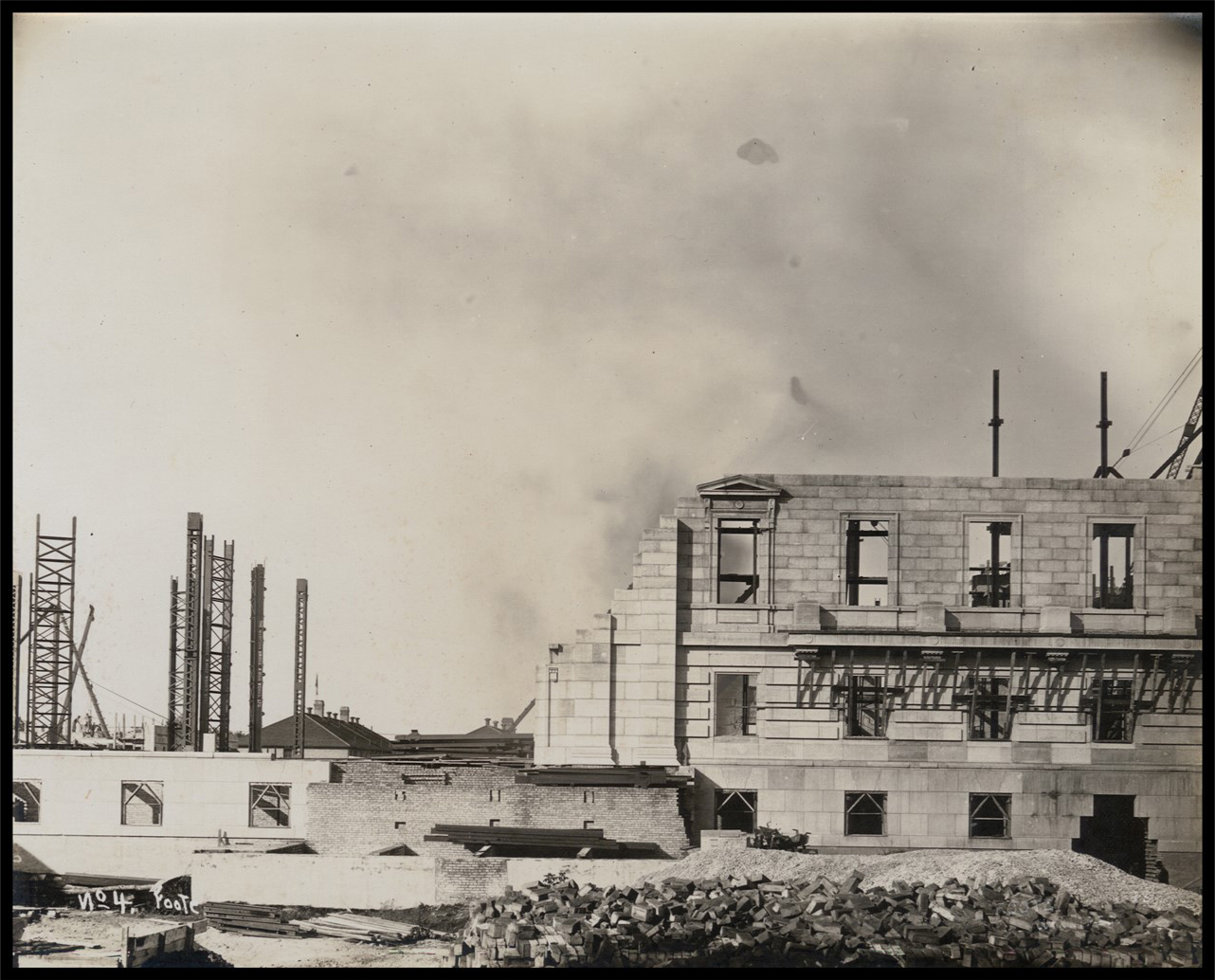 Photo of a half completed wall, with a pile of bricks in the foreground and steel pillars and buildings in the background.