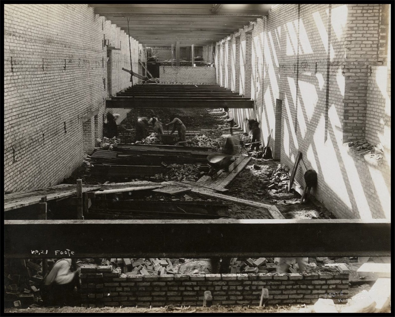 Photo of an interior portion of the building. The area is cluttered with construction materials. Large steel beams are suspended parallel to the floor, while workers build brick walls and push wheel barrows along temporary wood boardwalks.