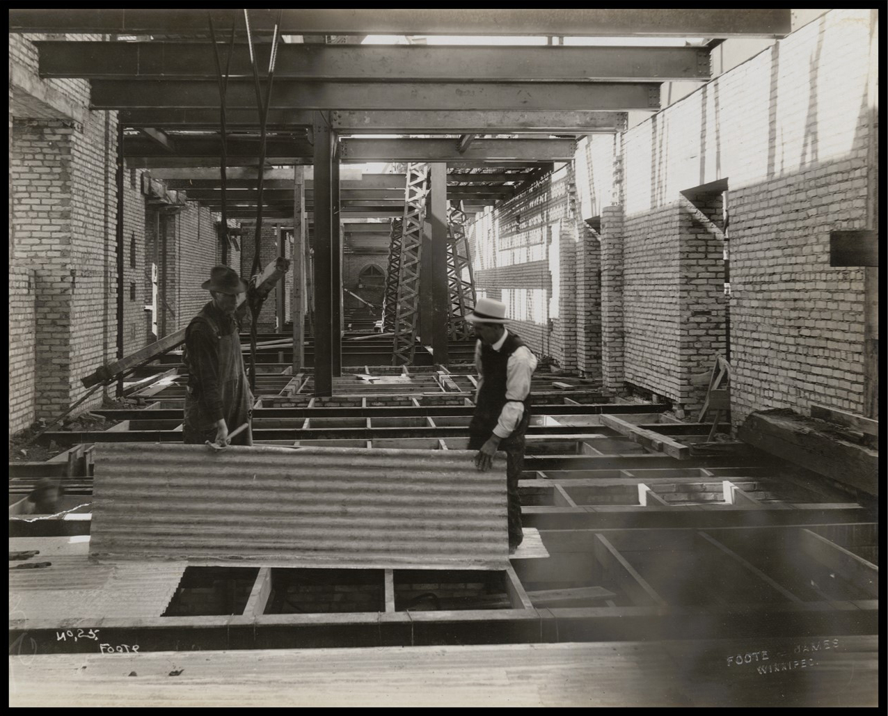 Photo of the interior of the building. The walls are complete, but the floors are still wood frames. Steel beams are exposed on the ceiling. Two men carry a large sheet to lay on the floor on top of the wood frames.