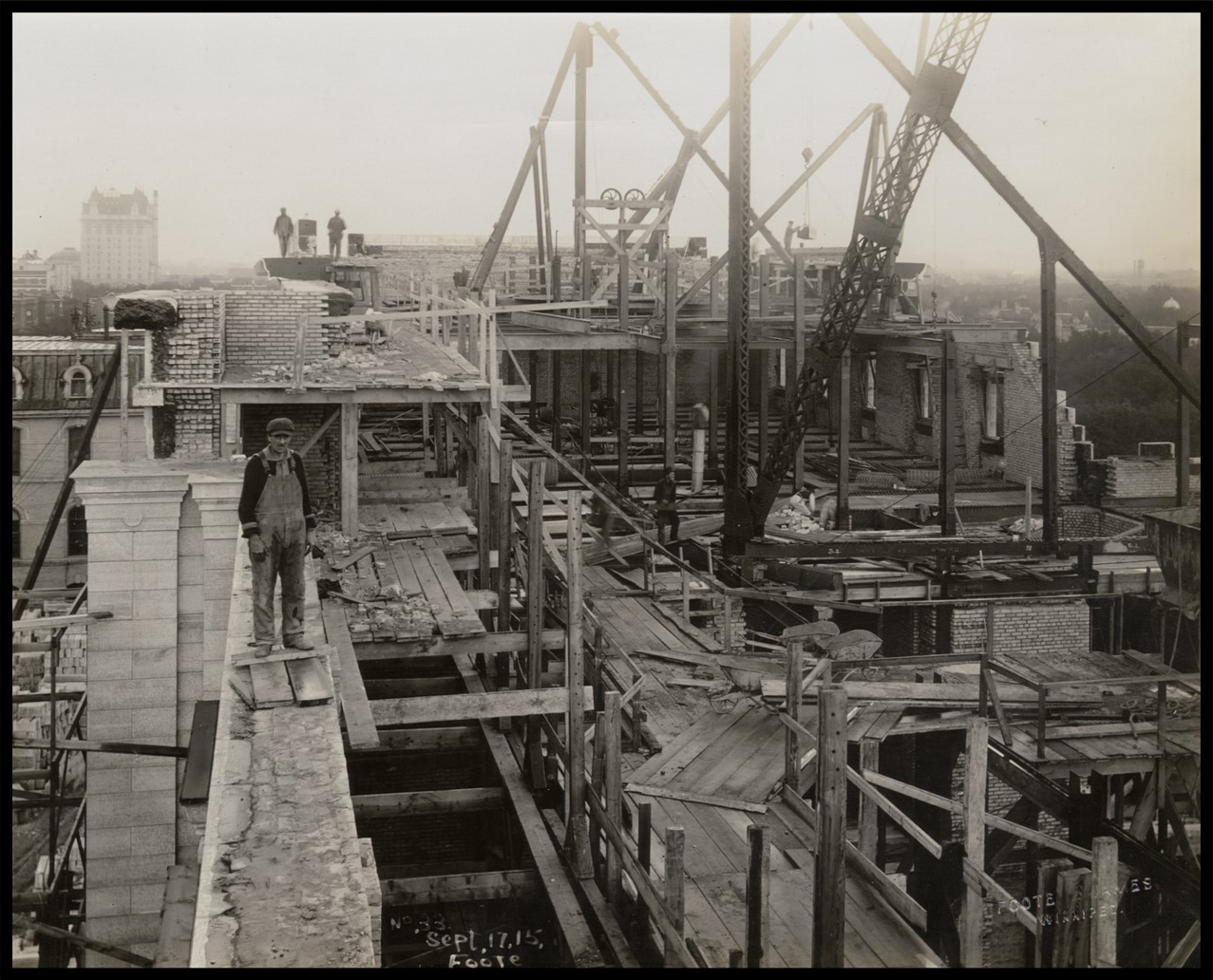 Photo of the top of the building under construction. The floors are temporary wood planks with protruding steel frames. Some of the brick walls are only partially complete. Several men stand throughout the area. One man is prominent in the foreground, standing on a plank. Other men sit on steel beams in the middle of the building. Two men stand on top of the wall in the distance. A man in the distance reaches towards a beam suspended by a crane.