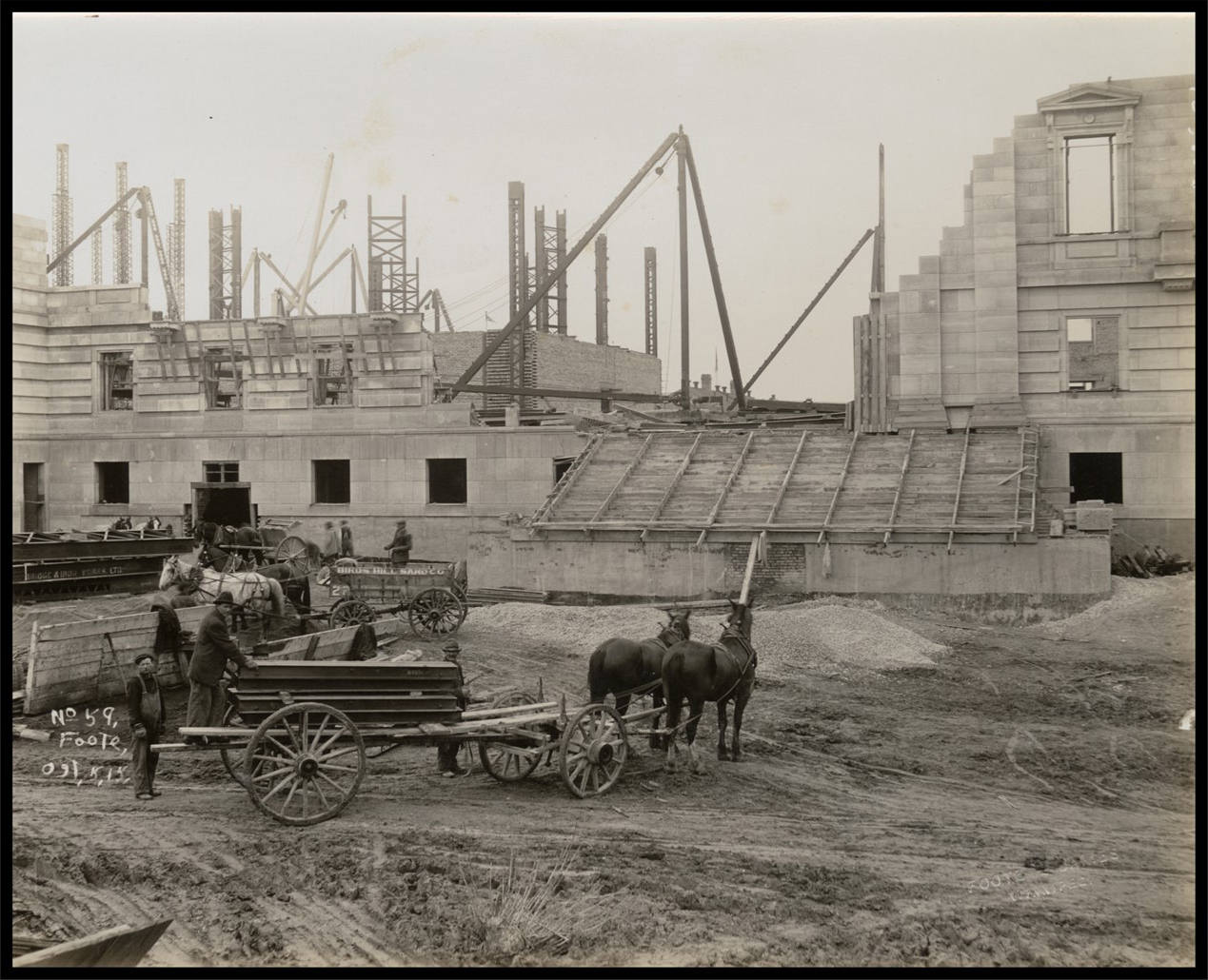 Photo of the back of the Legislative Building, with the walls only partially complete. Several men unload materials from horse drawn carriages.