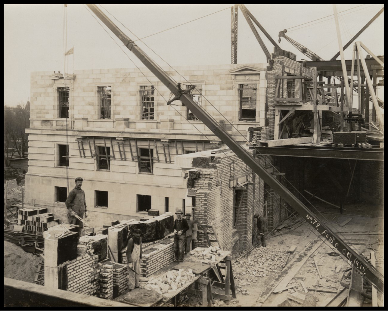 Photo from inside the building, looking down on several masons laying bricks for an exterior wall. A complete exterior wall can be seen through the incomplete brick wall.