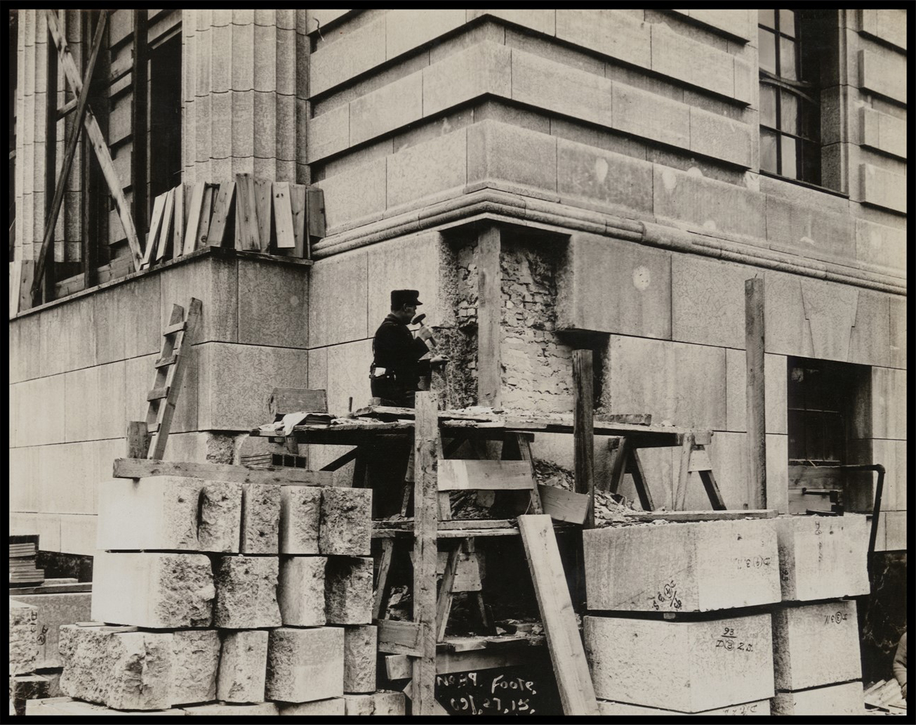 A mason working on large slabs of stone on the exterior base of the building.