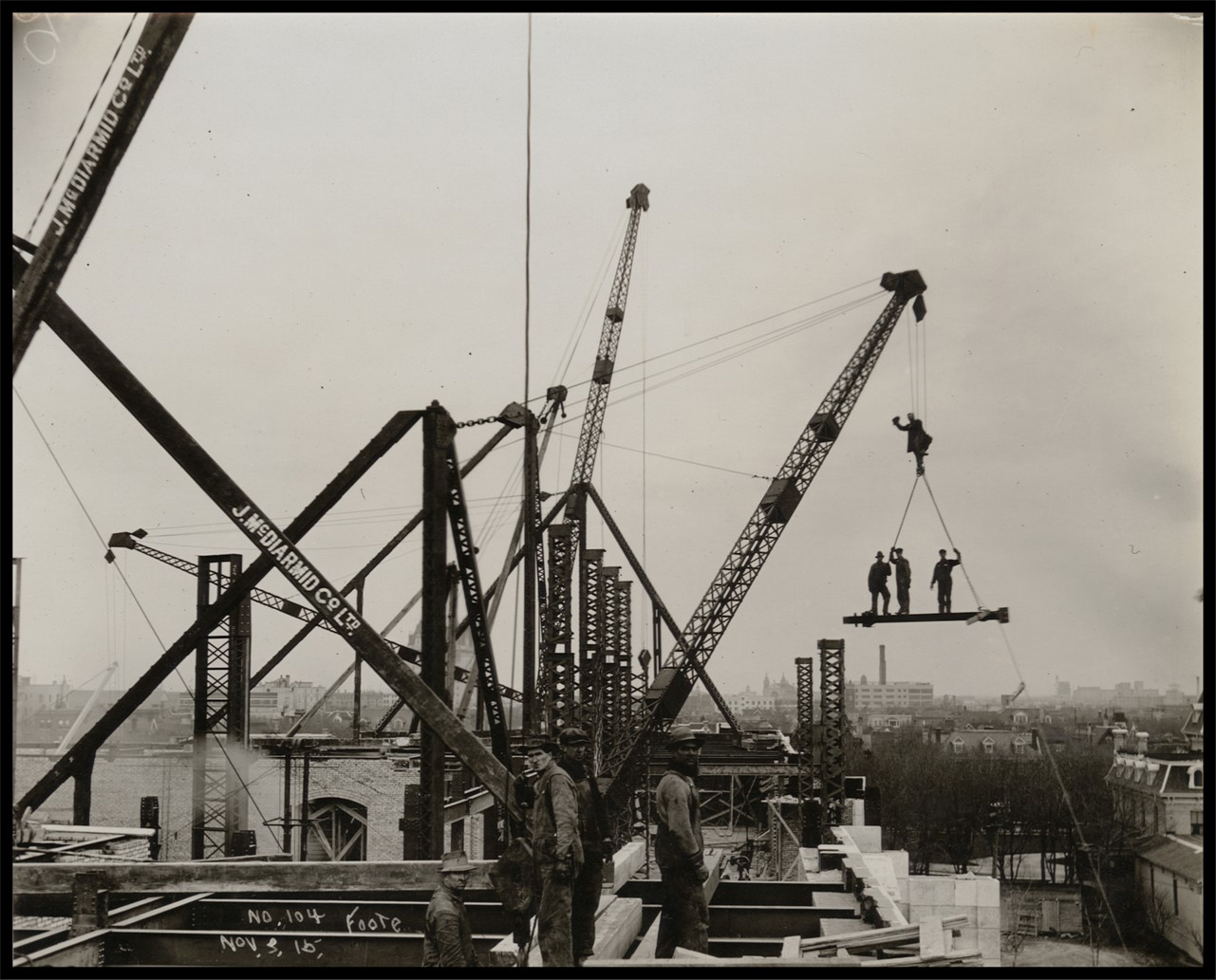 Photo of large cranes reaching up to the sky. Several men stand on the incomplete roof. Three men wave while standing on a beam suspended in mid-air, while another hangs onto the cable above them.