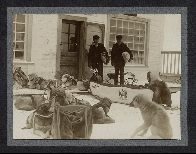 photo of two men outside an HBC post, holding bundles, and standing behind a dog sled and dogs