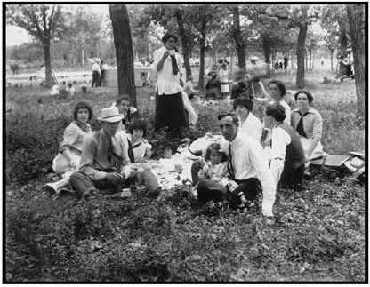 men, women, and children sitting on blanket in grass with food