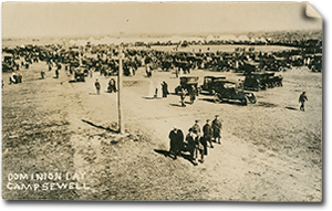 Front of postcard with photo of Camp Sewell on Dominion Day, 1915. There is a large group of people milling about in a field with parked cars. Many of the people are in uniform. In the distance, there is an open exhibition field with tents in the background.