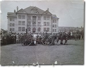historical photo of a large group of women standing in front of a building, watching as a man in uniform drives a horse drawn field kitchen on a cart. Two young boys in uniform and military officials also watch. A group of military soldiers stand at attention in the background.