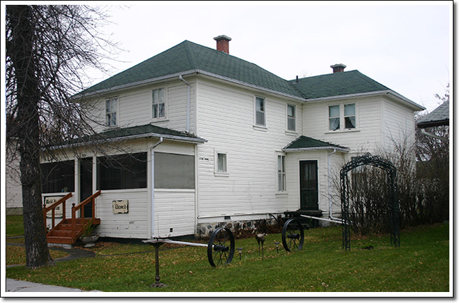 A white painted, two-storey frame house on an L-shaped plan, with a hipped roof and screened front porch