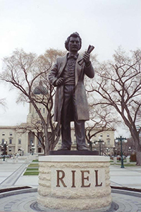 Statue en bronze de la figure complète de Louis Riel tenant un document dans son poing. La figurine est sur un piédestal qui indique RIEL.