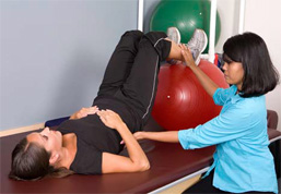 woman on her back with her legs on a yoga ball, another woman is assisting her